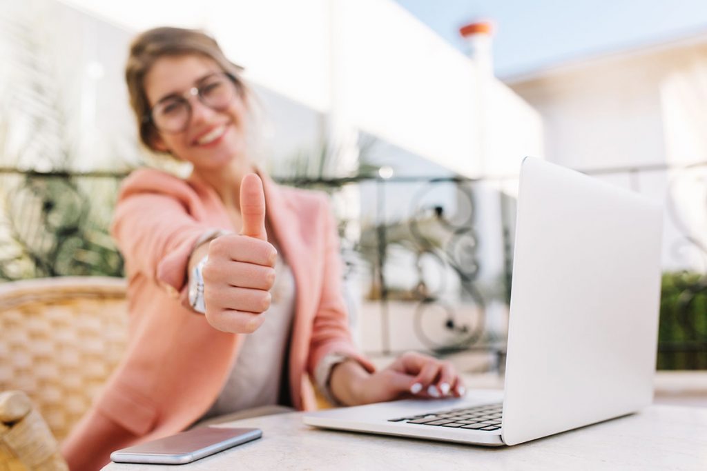 Cute young woman, student, business lady showing thumbs up, well done, sitting in outdoor cafe on terrace with laptop. Wearing pink smart clothes. .